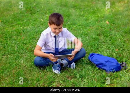 Un bon élève dans une chemise blanche et une cravate bleue sur l'herbe verte et regarde dans une tablette Banque D'Images