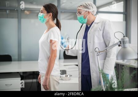 Un jeune médecin dans un masque effectue l'auscultation des poumons à partir de l'arrière de sa patiente. Banque D'Images