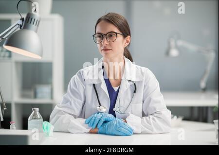 Une jeune interniste est assise à un bureau dans son bureau spacieux et lumineux, portant des gants et une blouse médicale. Banque D'Images