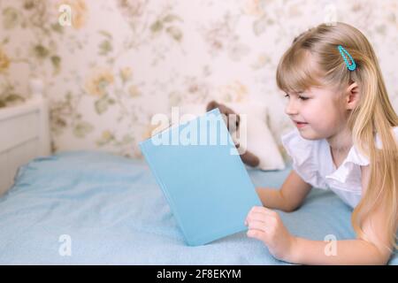 Une petite fille s'est allée sur le lit dans la chambre élégante et lit un livre bleu, faisant des devoirs. Éducation, concept de scolarisation à domicile Banque D'Images