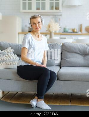 Portrait d'une femme âgée regardant un appareil photo assis sur un canapé dans un casque écouter de la musique dans le salon. Détendez-vous après avoir fait de l'exercice. Santé émotionnelle et spirituelle. Bien-être, bien-être pour femme à la retraite Banque D'Images