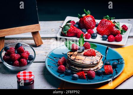 Dessert délicieux, crêpes au sirop de chocolat noir et fruits frais avec menthe sur une assiette bleue et une table en bois. Fond noir pour l'espace de copie Banque D'Images