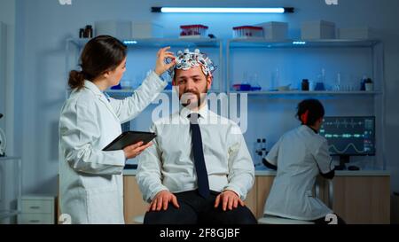 Jeune homme portant un casque de balayage d'onde de cerveau discutant avec un chercheur neurologique des symptômes de la maladie. Un scientifique médical écrit des informations sur le scan du cerveau sur une tablette travaillant en laboratoire Banque D'Images