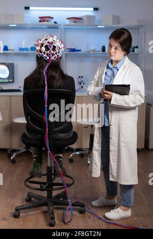 Une patiente porte un casque eeg performant qui balaie l'activité électrique du cerveau dans un laboratoire de recherche neurologique, tandis qu'une chercheuse médicale l'ajuste en examinant le système nerveux dactylographiant sur une tablette. Banque D'Images