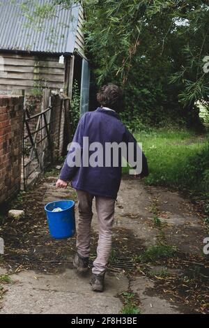 Vue arrière d'une femme sur une ferme, portant un seau en plastique bleu. Banque D'Images