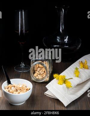 Un bol blanc contenant des arachides et d'autres fruits séchés à proximité récipients en verre avec vin noir et fleurs jaunes de sédum palmeri Banque D'Images