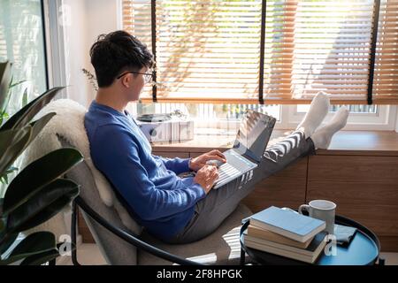 Jeune homme asiatique de technologie se détendant tenant un ordinateur portable et regardant l'écran dans le salon, travail à distance ou le concept de travail à domicile Banque D'Images