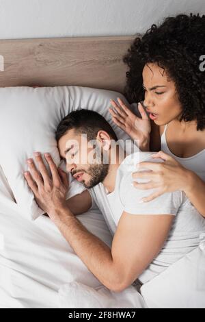 vue de dessus d'une femme afro-américaine mécontentée regardant barbu homme dormant sur le lit Banque D'Images