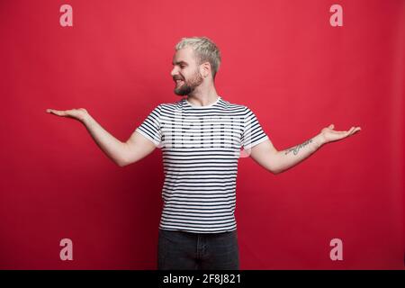 L'homme barbu caucasien sourit tout en tenant quelque chose et en comparant dans les palmiers posant sur un mur rouge foncé de studio Banque D'Images