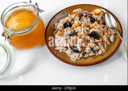 Porridge de flocons d'avoine avec morceaux de pomme, prunes séchées et miel dans une assiette avec cuillère et bol en verre avec miel sur fond blanc. Un petit déjeuner sain et sucré. Banque D'Images