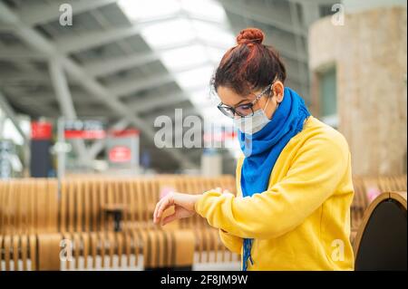 La femme caucasienne en masque utilise la montre intelligente sur le banc dans un lieu public. Banque D'Images