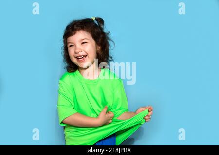 Photo d'une fille drôle de 4-5 ans en uniforme de sport rires sur fond bleu en studio, tirant vers le haut T-shirt. Regarde de côté Banque D'Images