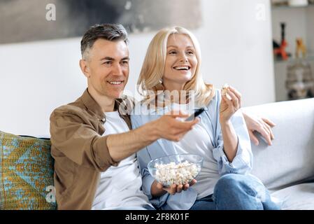 Regarder un film ensemble. Couple caucasien marié et heureux d'âge moyen assis à la maison sur un canapé, regarder la télévision, manger du pop-corn, regarder un film préféré, passer du temps libre à la maison, heureux ensemble, sourire Banque D'Images