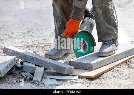 Un constructeur tient une scie électrique de travail avec une lame de diamant et coupe des blocs de granit dans l'arrière-plan du lieu de travail. Gros plan, arrière-plan flou. Banque D'Images