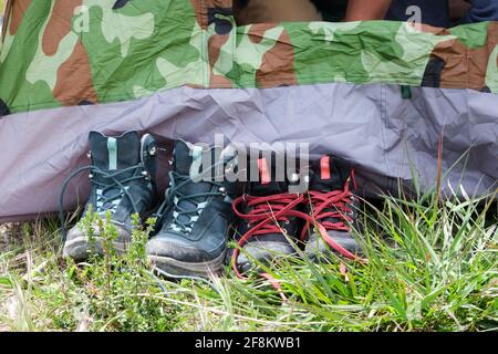 Deux paires de bottes de randonnée ou de marche pour enfants laissées bien alignées à l'extérieur d'une tente de camping camouflée sur un terrain d'herbe. Banque D'Images