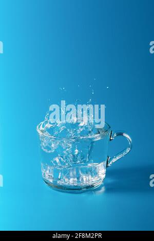 Retirez l'eau froide dans une tasse en verre sur fond bleu. Projection de glace tombant Banque D'Images