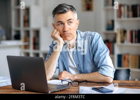 Homme d'affaires caucasien d'âge moyen ou indépendant assis sur le lieu de travail, travaillant sur un ordinateur portable, se reposant la tête sur la main à cause de la fatigue, faisant des heures supplémentaires, stressé et ennuyé, besoin de repos, regarder pensive loin Banque D'Images