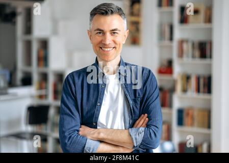 Portrait d'un homme, d'un indépendant ou d'un Manager caucasien aux cheveux gris mûrs, debout près de son bureau en chemise en denim avec les bras croisés, regardant et souriant amical à l'appareil photo Banque D'Images