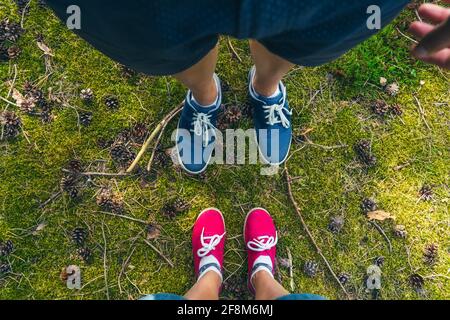 Regardez l'homme et la femme dans les sneakers de couleur qui se tiennent sur l'herbe verte avec des cônes et des branches d'arbre. Couple marchant dans la forêt d'été. Local Banque D'Images