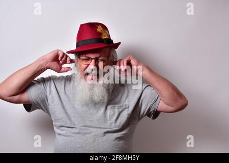 Vieux homme avec une chemise grise et un chapeau fedora rouge en bouchant ses oreilles avec ses doigts Banque D'Images