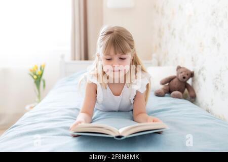 Une petite fille s'est allée sur le lit dans la chambre élégante et lit un livre bleu, faisant des devoirs. Éducation, concept de scolarisation à domicile. Tulipes jaunes dans le va Banque D'Images