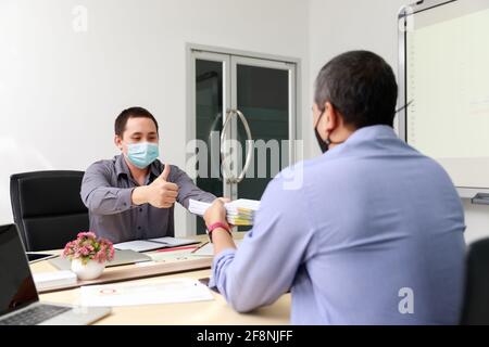 Les hommes d'affaires portant un masque de protection discutent et travaillent ensemble dans la salle de réunion. La pratique sociale à distance prévient le coronavirus COVID-19. Banque D'Images