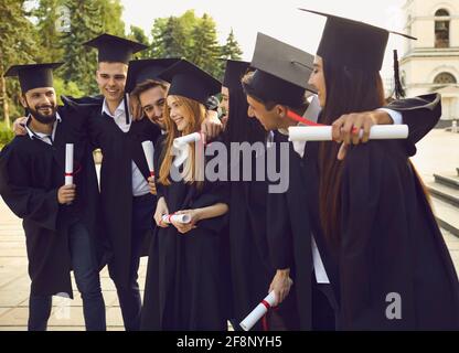 Groupe de jeunes diplômés universitaires souriants qui se tiennent debout avec des diplômes, s'embrassant Banque D'Images