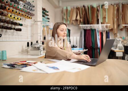 un designer de mode regarde l'ordinateur portable avec enthousiasme tout en travaillant avec des croquis dans un atelier d'atelier. Petite entreprise Banque D'Images
