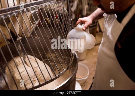 boulanger en gros plan plaçant de l'eau dans la préparation de pâte à pain à l'intérieur de la machine automatique de pétrissage de la boulangerie. conc. de cuisson industrielle automatisée Banque D'Images