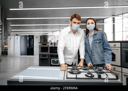 Jeune couple dans des masques médicaux regardant les appareils de cuisine dans un centre commercial Banque D'Images