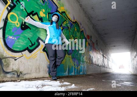 jeune femme en anorak bleu avec capuche et masque de protection se pencher contre un mur de graffiti dans un passage souterrain et avec ses bras s'étirent Banque D'Images