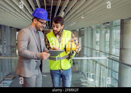 Architecte et ouvrier de construction debout dans le bâtiment en cours de construction et regardant les plans sur tablette. Banque D'Images