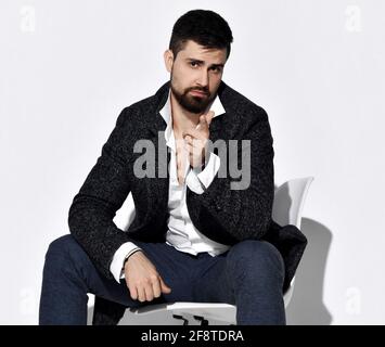 Jeune homme à barbe en Jean et veste en tweed décontractée est assis sur la chaise gestante pointant avec le doigt vers la caméra Banque D'Images