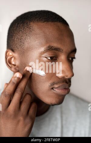 Jeune homme africain appliquant de la crème hydratante à sa joue Banque D'Images