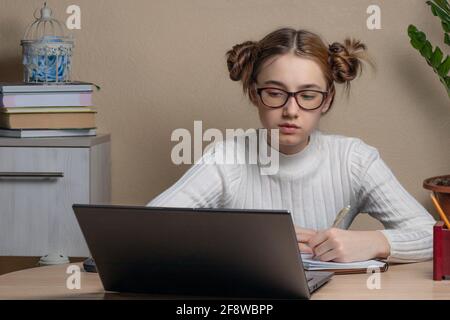 une jeune fille adolescente sérieuse autiful en lunettes est engagée en ligne sur un ordinateur portable. Une adolescente regarde un écran d'ordinateur portable et écrit quelque chose avec un stylo dans un non Banque D'Images