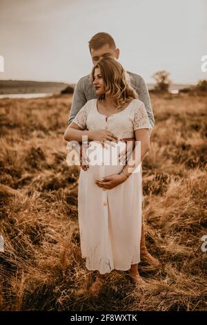 le mari se déchagine la femme enceinte garde ses mains sur un gros ventre rond. Une famille heureuse se reposant dans la nature embrasse les baisers en été au coucher du soleil. Future mère caucasienne Banque D'Images