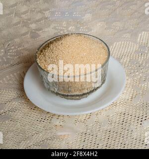 Une soucoupe blanche avec un plat crocory translucient rempli de sucre brun doré est sur une table recouverte d'une nappe recouverte d'ivoire coloré. Banque D'Images