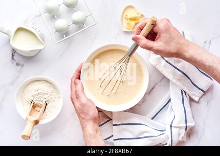 Les mains d'homme préparent la pâte pour les crêpes faites maison pour le petit déjeuner. Fouetter pour fouetter dans les mains. Ingrédients sur la table - farine de blé, oeufs, beurre, sucre, Banque D'Images