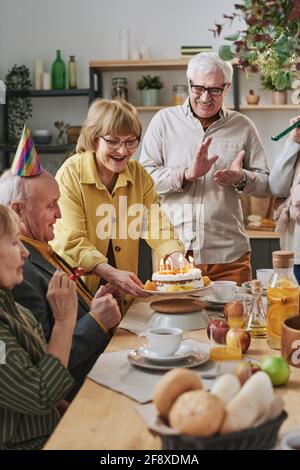 Groupe d'amis senior célébrant l'anniversaire avec un gâteau aux bougies tout en étant assis à la table de salle à manger ensemble à la maison Banque D'Images