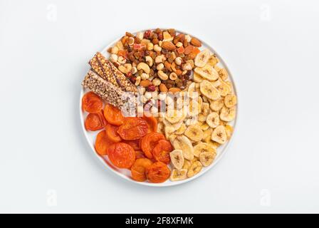 Barre de granola, abricots séchés, bananes séchées, noix et fruits secs dans une assiette plate blanche sur fond clair. Vue de dessus, horizontale. Une alimentation saine. Banque D'Images