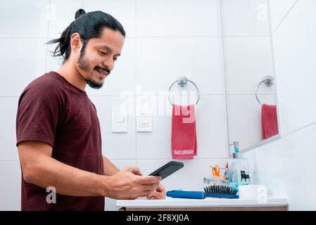 Jeune homme latin avec de longs cheveux noirs utilisant le téléphone tout en se brossant les dents dans la salle de bains Banque D'Images