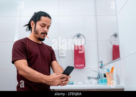 Jeune homme latin avec de longs cheveux noirs utilisant le téléphone tout en se brossant les dents dans la salle de bains Banque D'Images