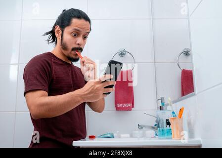 Jeune homme latin avec de longs cheveux noirs utilisant le téléphone tout en se brossant les dents dans la salle de bains Banque D'Images