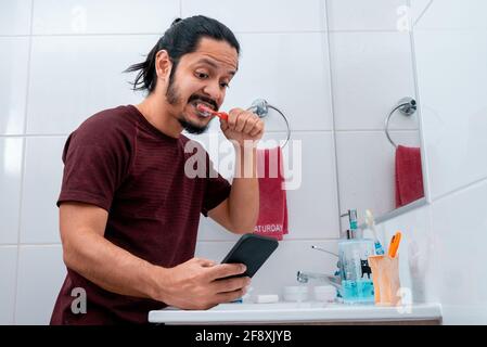 Jeune homme latin avec de longs cheveux noirs utilisant le téléphone tout en se brossant les dents dans la salle de bains Banque D'Images