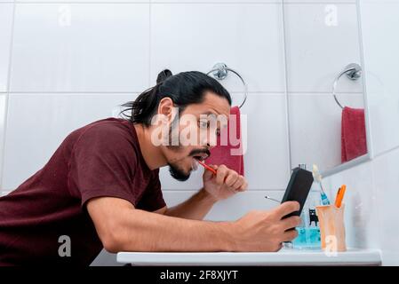 Jeune homme latin avec de longs cheveux noirs utilisant le téléphone tout en se brossant les dents dans la salle de bains Banque D'Images