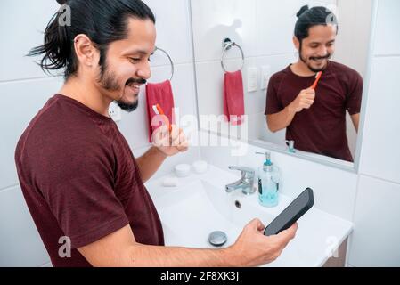 Jeune homme latin avec de longs cheveux noirs utilisant le téléphone tout en se brossant les dents dans la salle de bains Banque D'Images