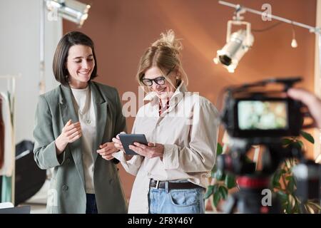 Femme d'affaires mûre riant tout en regardant quelque chose sur une tablette numérique avec sa collègue se tenant près d'elle au studio Banque D'Images