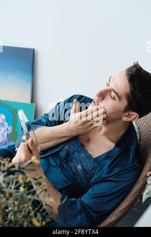 Jeune transgenre en robe féminine assis dans un fauteuil regardant miroir et soin de sa peau Banque D'Images