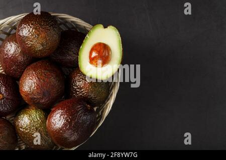 Avocats mûrs dans un panier sur une table noire, avec un fruit coupé et une pierre. Espace libre, vue de dessus Banque D'Images