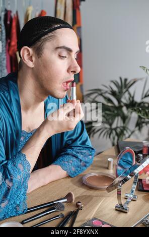 Jeune homme féminin assis à la table et appliquant du rouge rouge à lèvres sur ses lèvres Banque D'Images